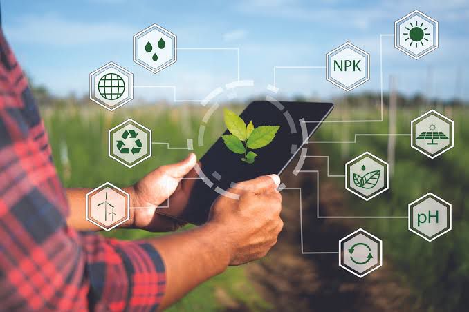 Um agricultor segura um tablet em meio a uma plantação, visualizando ícones digitais que representam indicadores ambientais, como pH do solo, nutrientes, energia renovável e reciclagem. A cena simboliza o uso da tecnologia e da inovação no agronegócio sustentável, destacando a integração entre práticas de cultivo responsáveis e soluções digitais voltadas para eficiência, produtividade e preservação ambiental.